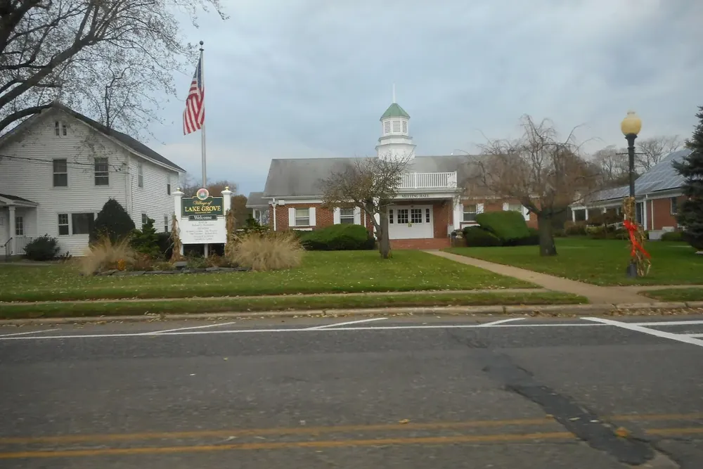 Lake Grove Village Hall with a flag and landscaped lawn beside the road in Lake Grove, New York