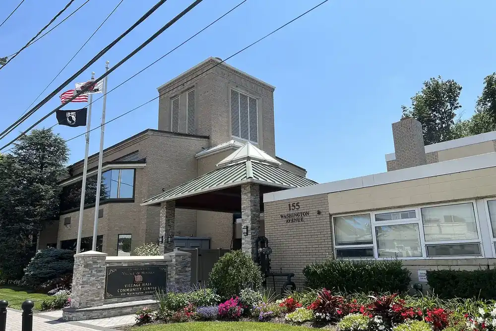 Mineola Village Hall with flags, a stone sign and bright flower beds in Mineola, New York